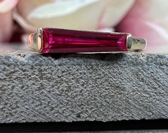 May include: A silver ring with a vibrant red rectangular gemstone. The ring is set on a gray surface.