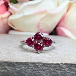 May include: A silver ring featuring a cluster of five round, deep red gemstones. The gemstones are arranged in a floral or starburst pattern. The ring is set against a wooden surface with a blurred floral background.