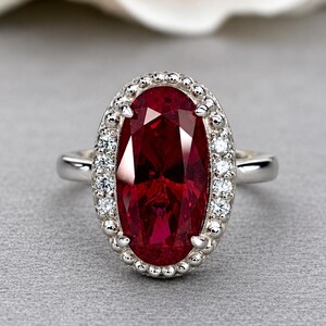 May include: A silver ring featuring a large, oval-cut, deep red gemstone. The gemstone is surrounded by a halo of small, clear gemstones. The ring's band is a simple, polished silver.