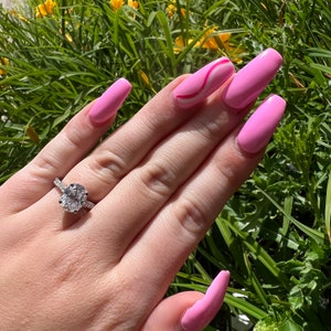 K&ouml;nnte beinhalten: Eine Hand mit rosa Nagellack und einem silbernen Ring mit einem gro&szlig;en ovalen Diamanten. Die Hand liegt auf einem Bett aus gr&uuml;nem Gras.