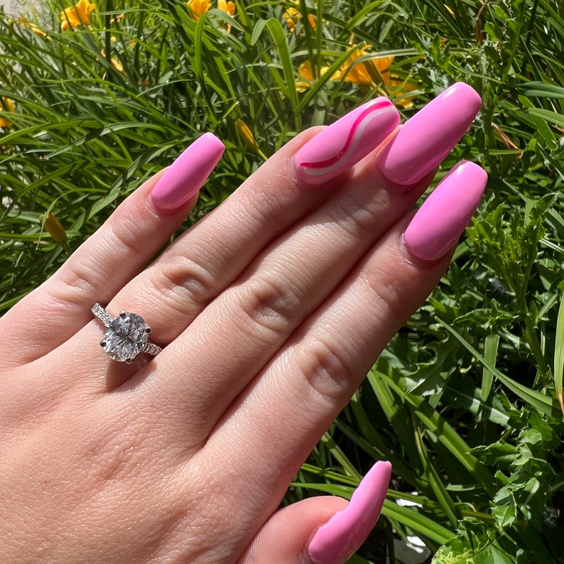 K&ouml;nnte beinhalten: Eine Hand mit rosa Nagellack und einem silbernen Ring mit einem gro&szlig;en ovalen Diamanten. Die Hand liegt auf einem Bett aus gr&uuml;nem Gras.