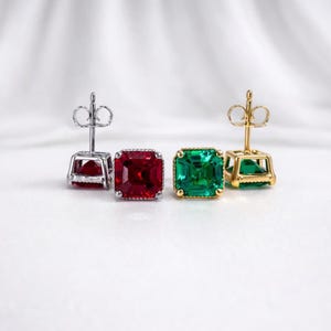 May include: Two pairs of square-cut gemstone stud earrings. One pair features a vibrant red gemstone set in a silver setting. The other pair showcases a bright green gemstone in a gold setting. The earrings are displayed on a white background.