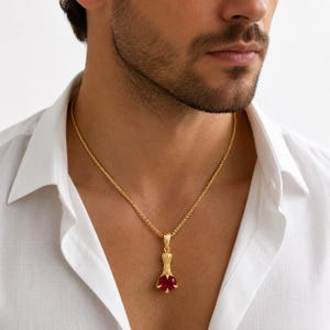 May include: A gold chain necklace featuring a pendant shaped like a bird's claw holding a round, faceted deep red gemstone. The pendant is suspended from a textured gold chain that rests against the collarbone of a person wearing a white shirt.