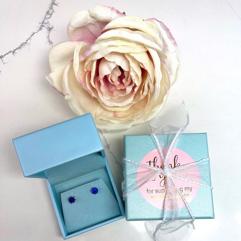 May include: A pair of blue sapphire stud earrings in a light blue gift box with a thank you tag. The tag has a pink circle with the text "Thank you for supporting my small business".