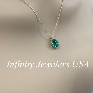 May include: A gold chain necklace with a green gemstone pendant surrounded by small diamonds. The pendant is in a prong setting. The necklace is on a mannequin's neck. Infinity Jewelers USA is written in white text at the bottom of the image.