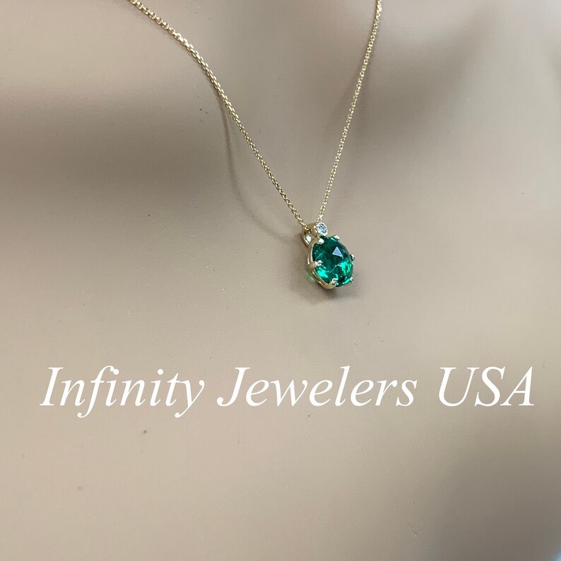 May include: A gold chain necklace with a green gemstone pendant surrounded by small diamonds. The pendant is in a prong setting. The necklace is on a mannequin's neck. Infinity Jewelers USA is written in white text at the bottom of the image.