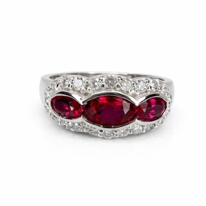 May include: A silver-toned ring featuring three oval-cut vibrant red gemstones arranged horizontally in the center, surrounded by a halo of small, clear, round-cut white stones set into the metal band that frames the red stones.