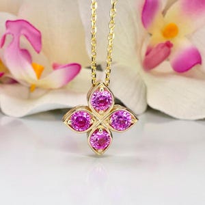 May include: A gold-colored pendant necklace featuring a four-petal flower design. Each petal holds a vibrant pink gemstone. The necklace hangs on a delicate gold chain, showcasing a piece of fine jewelry.