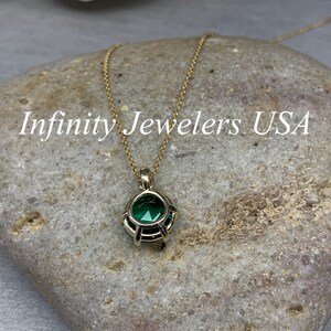 May include: A gold chain necklace with a green gemstone pendant. The pendant is set in a gold cage-like design. The necklace is on a gray rock with the text "Infinity Jewelers USA" in white.
