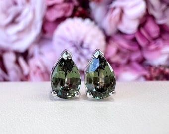 May include: A pair of teardrop-shaped earrings with a silver setting. The earrings feature a faceted, olive-green gemstone. The background is a soft focus of pink and purple flowers, creating a romantic aesthetic.