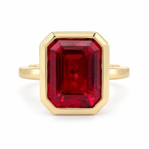 May include: A gold ring featuring a large, rectangular, emerald-cut red gemstone set in a smooth, polished gold bezel. The band is simple and rounded, providing a clean contrast to the prominent, vibrant red stone at the center of the piece.