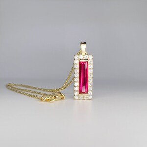 May include: A gold necklace with a rectangular pendant featuring a vibrant pink gemstone. The pendant is framed by a border of small, sparkling white gemstones. The chain and clasp are also gold.