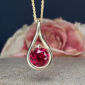 May include: A gold pendant necklace with a teardrop-shaped design. The pendant features a bright red gemstone in the center.