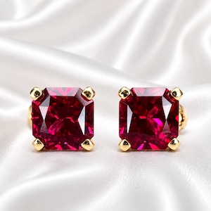 May include: A pair of stud earrings featuring square-cut, deep red gemstones set in polished yellow gold prongs. Each earring showcases a faceted surface that reflects light, highlighting the vibrant crimson color of the stones against the metal.