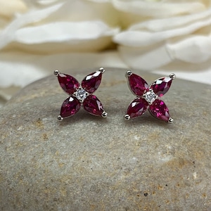 May include: A pair of silver stud earrings with a four-leaf clover design. Each clover is made of four red ruby stones and a single white diamond in the center.
