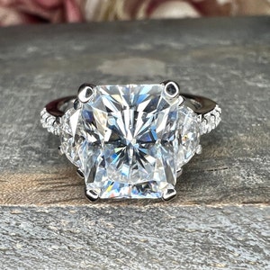 May include: A silver ring with a large, square-shaped, clear diamond in the center. The diamond is surrounded by smaller, clear diamonds on the band.