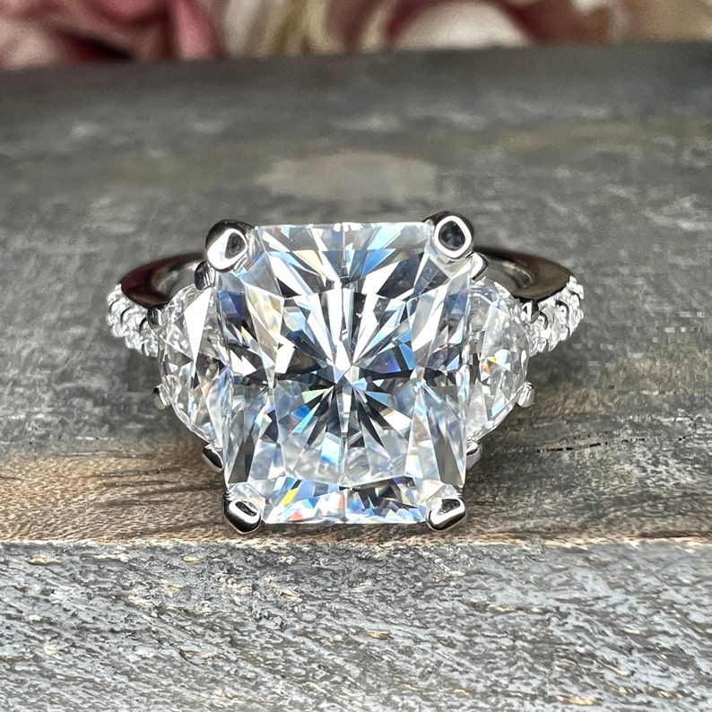 May include: A silver ring with a large, square-shaped, clear diamond in the center. The diamond is surrounded by smaller, clear diamonds on the band.
