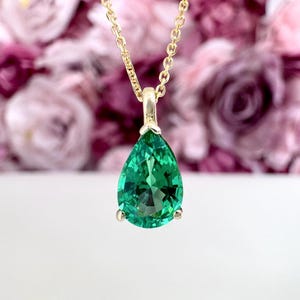 May include: A gold necklace with a pear-shaped, emerald-green gemstone pendant. The pendant is faceted, catching the light, and is suspended from a delicate gold chain. The background is a soft, blurred floral arrangement.
