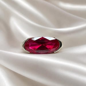 May include: A ring featuring a large, oval-cut, deep red gemstone set in a polished silver-toned metal band. The faceted stone displays a vibrant crimson hue with clear light reflections across its surface, held securely by two smooth metal prongs.