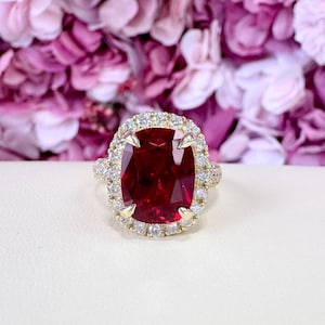May include: A gold ring featuring a large, cushion-cut, deep red gemstone surrounded by a halo of smaller, sparkling diamonds. The ring's band is also adorned with diamonds, creating a luxurious and eye-catching design.
