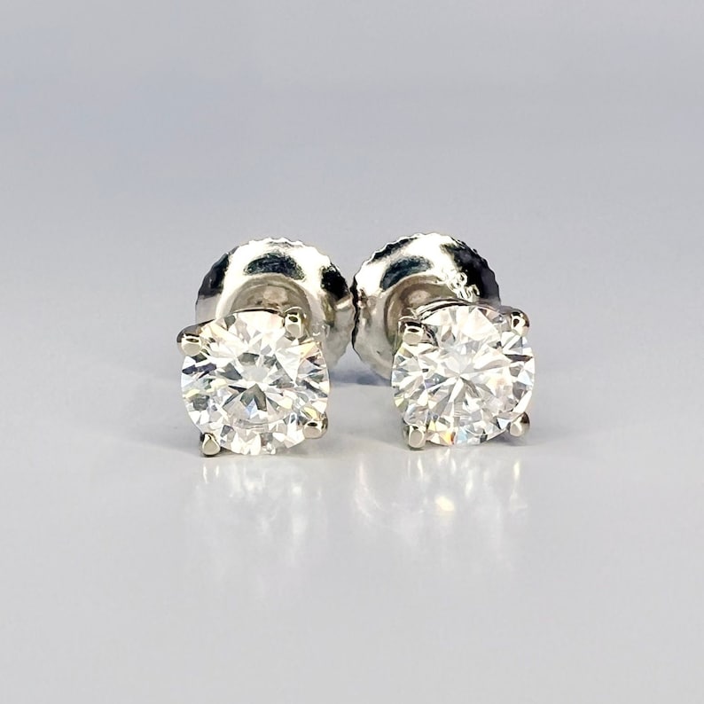 May include: A pair of diamond stud earrings. Each earring features a round, brilliant-cut diamond set in a four-prong setting. The diamonds sparkle brightly against a light background. The earring posts and backs are visible.