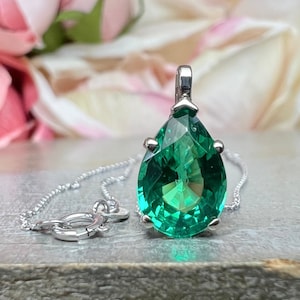 May include: A pear-shaped green gemstone pendant set in a silver-toned metal prong setting, suspended from a delicate silver-toned chain with a round spring ring clasp. The faceted stone displays a vibrant emerald hue with internal light reflections.