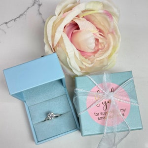 K&ouml;nnte beinhalten: Eine hellblaue Ringbox mit einem silbernen Ring mit einem klaren Stein darin. Die Box ist ge&ouml;ffnet und der Ring befindet sich in der Mitte der Box. Die Box steht auf einer wei&szlig;en Marmorfl&auml;che. Es gibt eine rosa und wei&szlig;e Geschenkbox mit einem wei&szlig;en Band und einem rosa Kreis mit dem Text "Vielen Dank f&uuml;r die Unterst&uuml;tzung meines kleinen Unternehmens" in goldenen Buchstaben.
