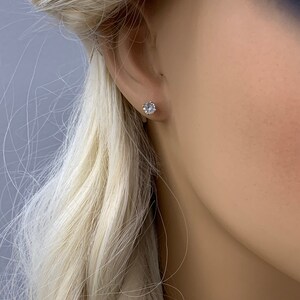 May include: A close-up of a person's ear with a small, round, clear gemstone stud earring.