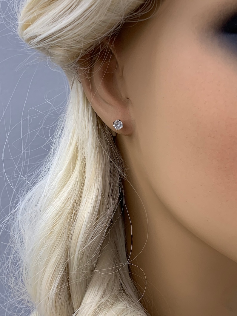 May include: A close-up of a person's ear with a small, round, clear gemstone stud earring.
