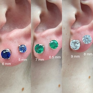 May include: A set of three pairs of stud earrings with different colored gemstones. The first pair features dark blue gems, 6 mm and 5 mm in diameter. The second pair has green gems, 7 mm and 6.5 mm. The third pair has clear gems, 9 mm and 8 mm.