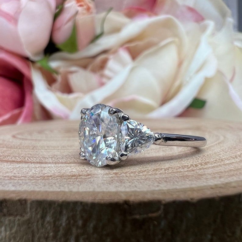 May include: A silver ring with a large oval-shaped center stone and two pear-shaped side stones. The ring is set on a wooden surface.