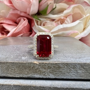 May include: A gold ring with a large, emerald-cut red gemstone surrounded by a halo of smaller, clear gemstones.