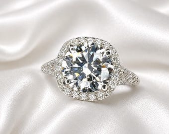 May include: A silver-toned ring featuring a large, round, brilliant-cut center stone held by four prongs. The center stone is surrounded by a square-shaped halo of small, round, pavé-set stones, with additional small stones lining the band.
