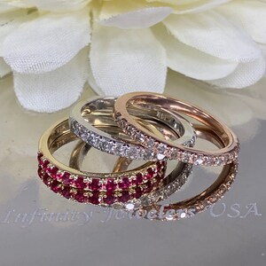 May include: Three eternity bands with a row of gemstones. The top band is rose gold with small diamonds, the middle band is white gold with small diamonds, and the bottom band is yellow gold with a row of red rubies.