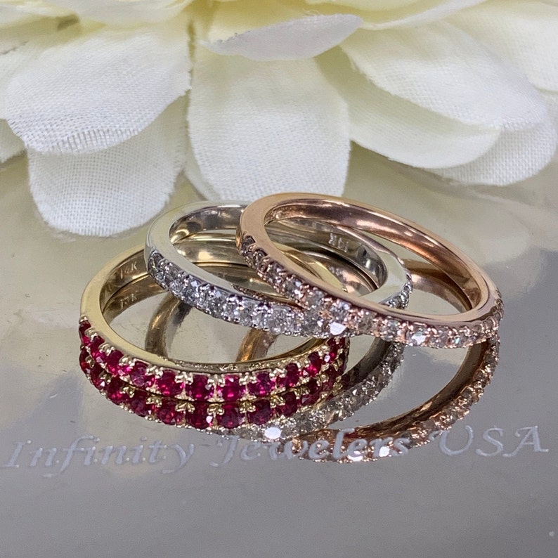May include: Three eternity bands with a row of gemstones. The top band is rose gold with small diamonds, the middle band is white gold with small diamonds, and the bottom band is yellow gold with a row of red rubies.