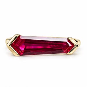 May include: A gold ring featuring a long, tapered, baguette-cut ruby gemstone set horizontally. The vibrant red stone is secured by gold prongs at both pointed ends, creating a sleek, elongated profile that spans across the finger band.