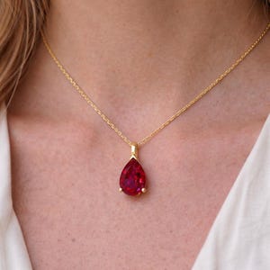 May include: A gold necklace featuring a pear-shaped, deep red gemstone pendant. The pendant is set in a gold bezel and hangs from a delicate gold chain. The necklace is displayed against a neutral background.