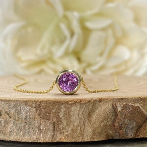 May include: A delicate gold chain necklace with a round bezel setting featuring a vibrant purple gemstone.