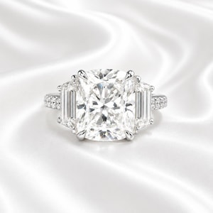 May include: A silver-toned ring featuring a large, central cushion-cut stone held by four prongs. The center stone is flanked by two rectangular baguette-cut stones on each side, with a band accented by small, round pavé-set stones along the sides.