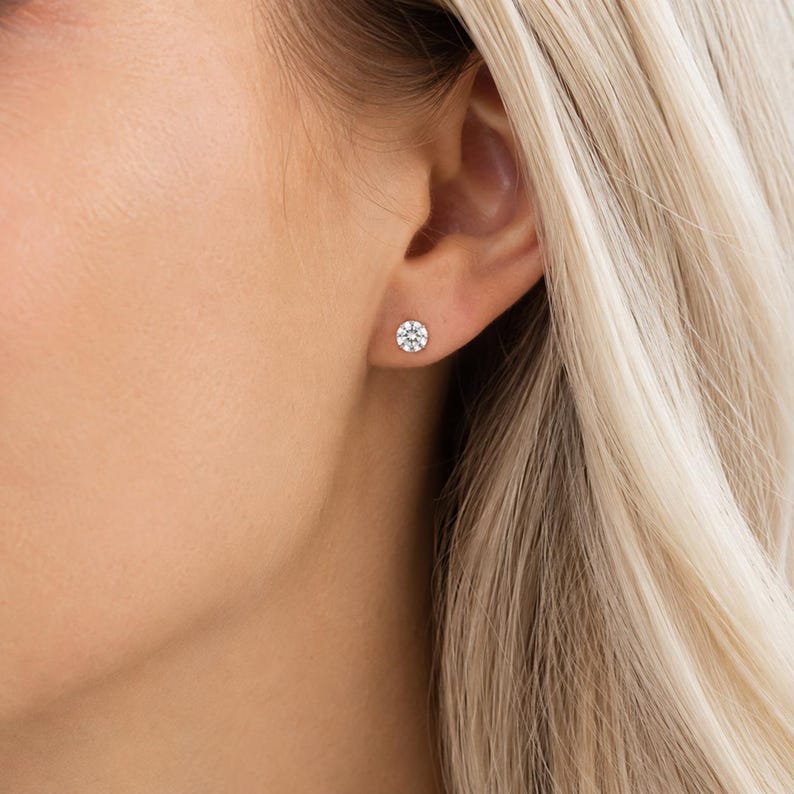 May include: A close-up of a woman's ear with a small, round stud earring. The earring is composed of multiple small, clear stones arranged in a circular pattern. The earring is set against the backdrop of the woman's skin and blonde hair.