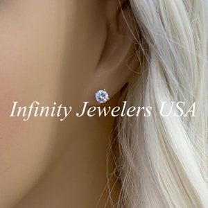 May include: A close-up of a sparkling, round-cut diamond stud earring. The earring is set in a silver or white gold setting and is worn on an ear. The text "Infinity Jewelers USA" is visible.