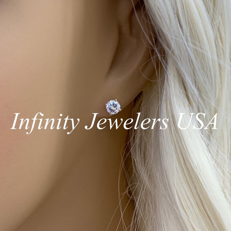 May include: A close-up of a sparkling, round-cut diamond stud earring. The earring is set in a silver or white gold setting and is worn on an ear. The text "Infinity Jewelers USA" is visible.