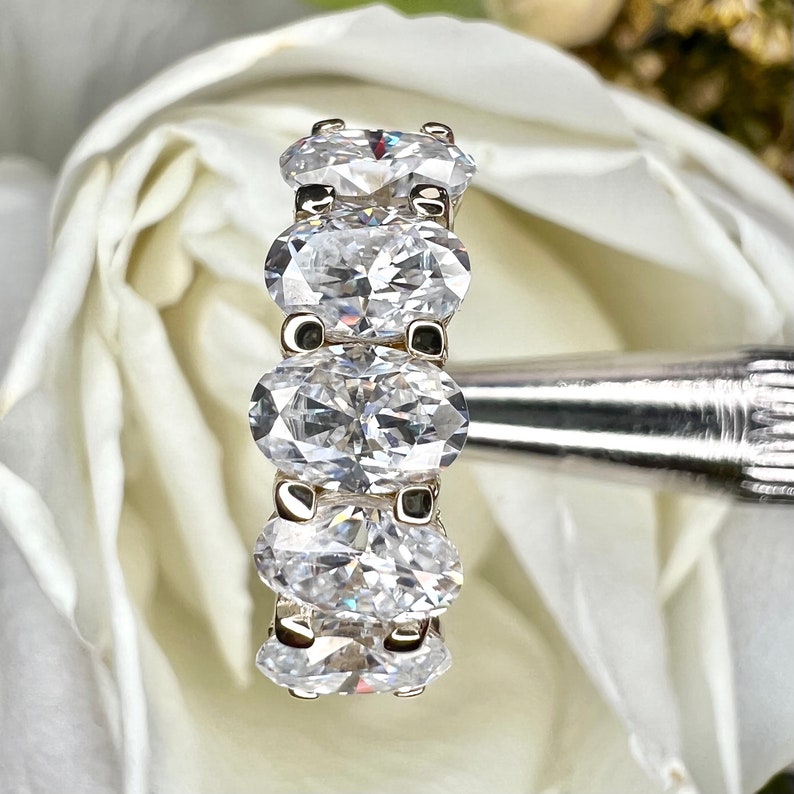 May include: A silver band ring with five oval-shaped clear gemstones. The ring is set against a white background.