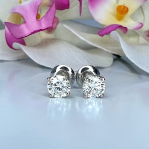 May include: A pair of diamond stud earrings with a four-prong setting. The round-cut diamonds sparkle brightly against a soft background, with a blurred orchid in the background. The earrings are set on a silver post.