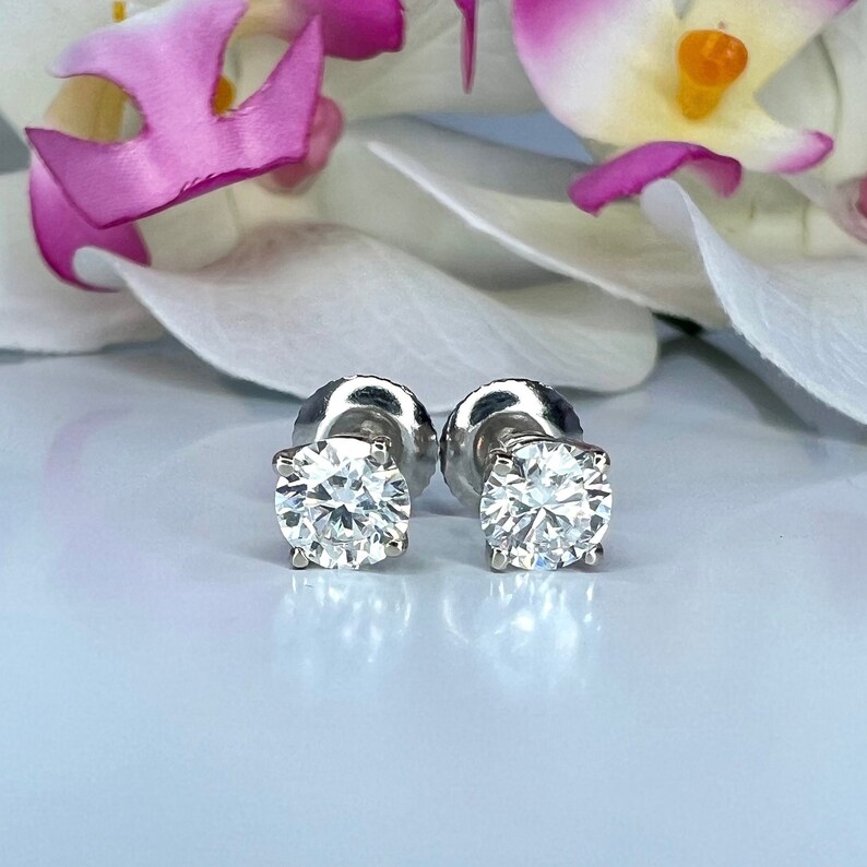 May include: A pair of diamond stud earrings with a four-prong setting. The round-cut diamonds sparkle brightly against a soft background, with a blurred orchid in the background. The earrings are set on a silver post.