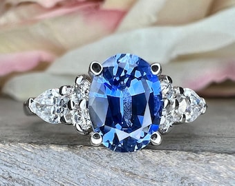 Op de afbeelding: Een zilveren ring met een grote ovale blauwe saffier, omringd door kleinere diamanten. De ring heeft een drie-klauw zetting en is versierd met peervormige diamanten aan weerszijden van de saffier.