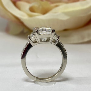 May include: A silver ring with a large square-shaped center stone and two smaller stones on either side. The ring has a band of small stones.