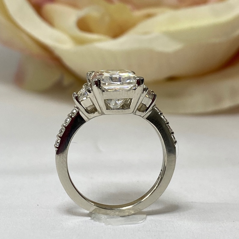 May include: A silver ring with a large square-shaped center stone and two smaller stones on either side. The ring has a band of small stones.