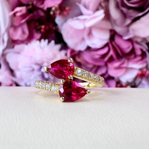 May include: A gold ring featuring two pear-shaped, vibrant red gemstones. The ring band is adorned with small, sparkling white stones. The background is a soft focus of pink and purple flowers, creating a romantic aesthetic.