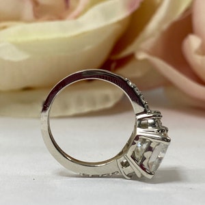 May include: A silver ring with a large, clear, square-shaped gemstone surrounded by smaller gemstones. The ring has a band that is set with smaller gemstones.
